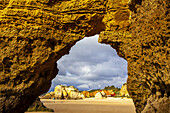 Looking through the detail of a sea arch on Dona Ana Beach at low tide with its sunlit shoreline and sandstone sea stacks along the famous Algarve coastline under a stormy sky; Praia da Dona Ana, Lagos, Algarve Region, Portugal