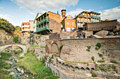 A small arch bridge crossing the Tsavkisi-Tskali River with love locks attached to its railings in front of the old buildings on the cliffs in Legvtakhevi, part of the historical neighborhood of Abanotubani in the Old Town with the domes of the hot sulfur bath houses above the retaining wall and the minaret of the Tbilisi Mosque in the background; Tbilisi, Georgia