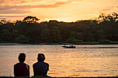 Blick von hinten auf eine Silhouette von Touristen, die am Ufer sitzen und einen goldenen Sonnenuntergang mit einem Boot beobachten, das die Wasserwege um Tortuguero befährt; Provinz Limon, Costa Rica