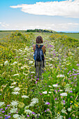View taken from behind of a woman standing in a wild flower meadow wearing a backpack, looking at the coastal view with Torre Sant'Emiliano in the distance near Otranto; Otranto, Puglia, Italy