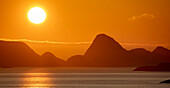 Mitternachtssonne leuchtet hell am orangefarbenen Himmel über den Berggipfeln und spiegelt sich im Nordatlantik; Lofoten, Polarkreis, Norwegen