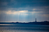 Hellisoy-Leuchtturm, ein abgelegener Leuchtturm auf einer Insel in den westlichen Fjorden Norwegens mit Sonnenstrahlen, die sich in einem stürmischen Himmel spiegeln; Hordaland, Norwegen