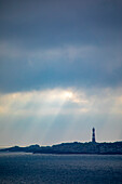 Hellisoy-Leuchtturm, ein abgelegener Leuchtturm auf einer Insel in den Westfjorden Norwegens mit Sonnenstrahlen, die sich in einem stürmischen Himmel spiegeln; Hordaland, Norwegen