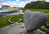 Die dramatische Küstenberg- und Felslandschaft des Uttakleiv-Strandes auf den Lofoten, Norwegen, mit dem natürlich geformten herzförmigen Felsen, der auf einem großen Felsblock balanciert, und anderen Felsen, die in einem herzförmigen Muster auf dem Gras liegen; Lofoten, Norwegen.