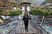 Eine Frau steht auf einer mit Gebetsfahnen gesäumten Hängebrücke aus Eisen; Tamchhog Lhakhang Bhutan