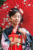 Porträt eines jungen Mädchens, das einen Kimono trägt und einen roten Papierschirm hält; Tokio, Japan.