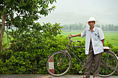 Ein Mann steht mit seinem Fahrrad neben einem üppigen grünen Feld; Mangshi Yunnan China
