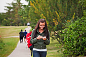 Junge Frau, die auf einem Pfad in einem Park spazieren geht und Textnachrichten abruft; Edmonton Alberta Kanada
