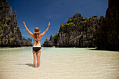 A Woman Tourist Wearing A Sun Hat And Bikini Stands With Arms Raised In The Clear Waters Of Matinloc Island Near El Nido And Corong Corong; Bacuit Archipelago; Palawan Philippines