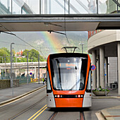 Public Transportation Train On A City Street With A Walkway Overhead And A Rainbow In The Background; Bergen Norway