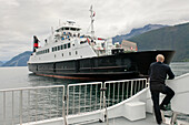Ein Mann steht am Heck eines Bootes und beobachtet ein vorbeifahrendes Schiff; Sognefjord Norwegen