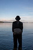 Mann in Anzug und Hut hält einen Felsbrocken, während er in einem ruhigen See bei Sonnenuntergang steht