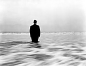 Rückansicht eines mittelgroßen erwachsenen Mannes in Anzug und Hut, der knietief im Meerwasser steht