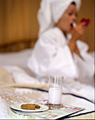 Junge erwachsene Frau im Bademantel trägt Lippenstift im Hotelzimmer auf, im Vordergrund Milch und Kekse