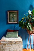 Chest with cushions, houseplant and painting on blue wall in bedroom