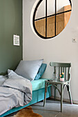 Schlafzimmer mit Einzelbett, rundem Fenster, grüner Wand und grünem Holzstuhl als Nachttisch