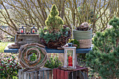 Winterdeko mit Fichtenbäumchen, Kiefernzweigen, Herbstkranz, Zapfen auf Gartentisch und Windlicht auf Kiste