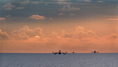 Fischerboote tief draußen auf dem Meer vor dem Hintergrund dramatischer Wolken und des Himmels bei Sonnenuntergang.