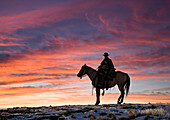 USA, Shell, Wyoming. Hideout Ranch cowgirl silhouetted on horseback at sunset. (PR,MR)