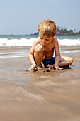 Ein Junge im Urlaub spielt mit Sand am Strand von Patnum, Goa, Indien.
