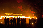 Silhouttes Of People Watching Procession Of Bonfire Societies With Burning Torches Coming Down Country Lane With Sky Lit By Red Flares On Bonfire Night, Barcombe, East Sussex, Uk