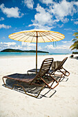 Liegestühle unter Sonnenschirm am weißen Sandstrand mit Palmen und Blick auf das blaue Meer. Pantai Cenang (Cenang Beach), Pulau Langkawi, &#10; Malaysia, Südostasien.