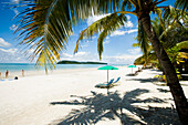 Malaysia, Pantai Cenang (Cenang Strand); Pulau Langkawi, Liegestühle unter Sonnenschirm am weißen Sandstrand mit Palmen mit Blick auf das blaue Meer