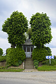 Stufen, die zu einer kleinen Kapelle führen, umgeben von Kastanienbäumen mit einem blau-weißen Chevron-Schild an einer Straße bei Arras; Nordfrankreich, Frankreich