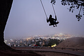 At dusk, a woman swings under a tree at Bernal Heights over San Francisco fog.; San Francisco, California, United States of America