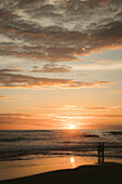 Silhouette von zwei Menschen, die am Wasser stehen und den Sonnenuntergang am Strand beobachten; Munggu, Bali, Indonesien
