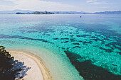 Aerial view of the edge of a white sand beach and the turquoise water along an island with boats moored offshore and the mountain ridges of islands on the horizon in Komodo National Park; East Nusa Tenggara, Lesser Sunda Islands, Indonesia