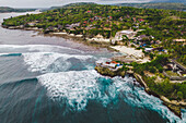 Luftaufnahme der Meeresbrandung und der rollenden Wellen entlang der Strandpromenade des Ferienortes Lembongan mit üppiger Vegetation und Gebäuden am Rande des türkisfarbenen Wassers vor der Küste Balis; Nusa Islands, Klungkung Regency, Ost Bali, Indonesien