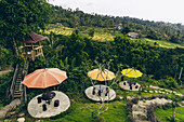 Lookout tree and tables with colorful umbrellas looking out at the rice fields and farm buildings on the hillside in Sambangan in the Sukasada District; Buleleng, Bali, Indonesia