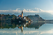 Blick über das ruhige Wasser des Bleder Sees in der Morgendämmerung auf die Kirche auf der Bleder Insel und das Schloss Bled dahinter; Bled, Slowenien