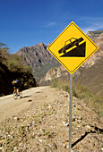 A biker heads downhill past a steep slope sign.; Batopillas Canyon, Sierra Madre Mountains, Mexico.