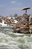 A stand up paddle boarder poses on rocks below Great Falls.; Potomac River, Maryland.