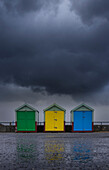 Three brightly colored beach huts sit beneath a storm cloud on a wet autumnal day; Hove, East Sussex, England, United Kingdom