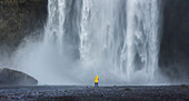 A lone tourist stands in front of the impressive Skogafoss waterfall in Iceland as they prepare to take a photo; Skogafoss, Iceland
