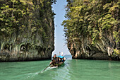 Traditional, Thai longtail boat traveling through the green, turquoise colored waters in between too vast, karst rock formations; Phang Nga Bay, Thailand