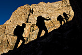 Vor dem Hintergrund der sonnenbeschienenen Klippen klettert das vierköpfige Team die größte aller Geröllhalden hinauf, auf die wir gestoßen sind, bis zu einer dritten Ebene an der nach Nordwesten ausgerichteten Klippenwand, um zwei große Höhleneingänge zu erkunden, die als zu gefährlich für den Zugang ohne Seil gemeldet worden waren.