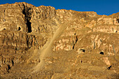 As the evening sunlight arrives, an expedition team member stands upon a large lump of rock. In the distance and beyond him, another team member stands in blue in the middle of the righthand entrance to The U-shaped cave. The giant scree slope, the largest of all the scree slopes of rocks the team endured on this expedition drops away in the middle of the cliff face, providing the access they needed to get into The Crystal Palace.