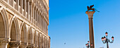 Die geflügelte Löwenstatue auf der Markus-Säule auf dem Markusplatz mit den weißen Bögen des Dogenpalastes (Palazzo Ducale) vor blauem Himmel; Veneto, Venedig, Italien