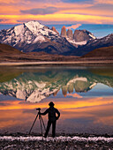 Blick von hinten auf die Silhouette eines Fotografen mit Kamerastativ am Ufer des Lago Azul bei Sonnenaufgang auf der Jagd nach Pumas; Torres del Paine National Park, Patagonien, Chile
