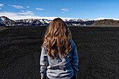Close-up view of a woman with blond hair, taken from behind, standing in front of a massive landscape of black sand with a mountain ridge and a blue sky in the distance; Iceland