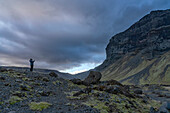 Woman standing out on the volcanic, rocky landscape admiring the natural beauty and taking a picture of the mountains and dramatic storm clouds forming overhead; South Iceland, Iceland