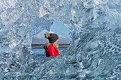 Nahaufnahme einer Frau mit Wollmütze, die durch eine runde, klare Eisformation hinausschaut und die Eisberge an der Südküste Islands bewundert; Jokulsarlon, Südisland, Island