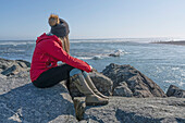 Blick auf eine Frau, die auf den Felsen am Strand sitzt und die Eisberge an der Südküste Islands vorbeischwimmen sieht; Jokulsarlon, Südisland, Island