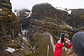 Close-up view, taken from behind, of a woman standing and overlooking Mulagljufur Canyon, a hikers paradise, taking a picture with her smart phone of an amazing view of a waterfall through the moss-covered cliffs; Vik, South Iceland, Iceland