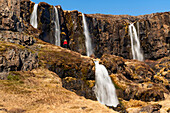 Scenic view of a woman standing on a cliff side slope in front of a series of waterfalls, some above and below, flowing from the craggy cliffs of the East Fjords making her appear small against the vast landscape in front of her; East Iceland, Iceland