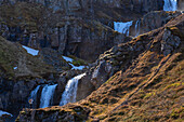 Landschaftliche Ansicht einer Frau, die ein Foto mit ihrem Smartphone macht, stehend auf einem Klippenhang vor einer Reihe von Wasserfällen, die von den zerklüfteten Klippen der Ostfjorde herabstürzen und sie gegen die weite Landschaft vor ihr klein erscheinen lassen; Ostisland, Island